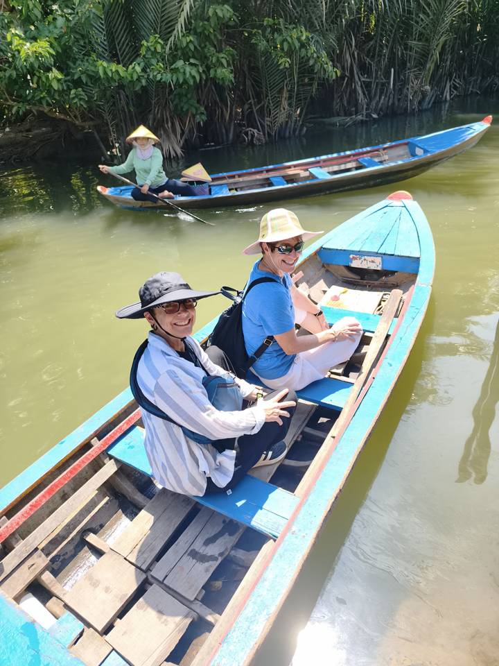 Two smiling women sit in wooden boat on calm green canal under bright sun.