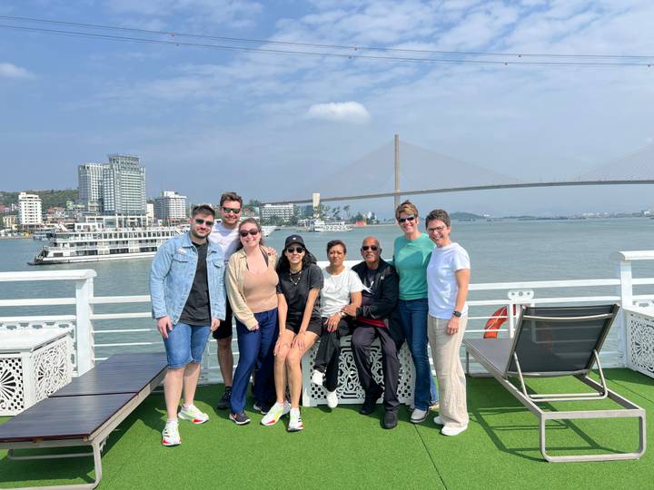 Group photo on deck of cruise ship in Halong Bay with bridge and city skyline behind.