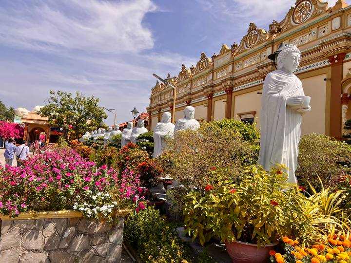 Colorful garden with blooming flowers and a row of white Buddhist statues at ornate temple complex.