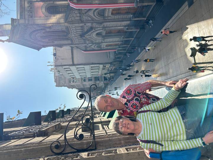 Couple poses on a balcony overlooking a pedestrian street lined with historic buildings under bright sunlight.