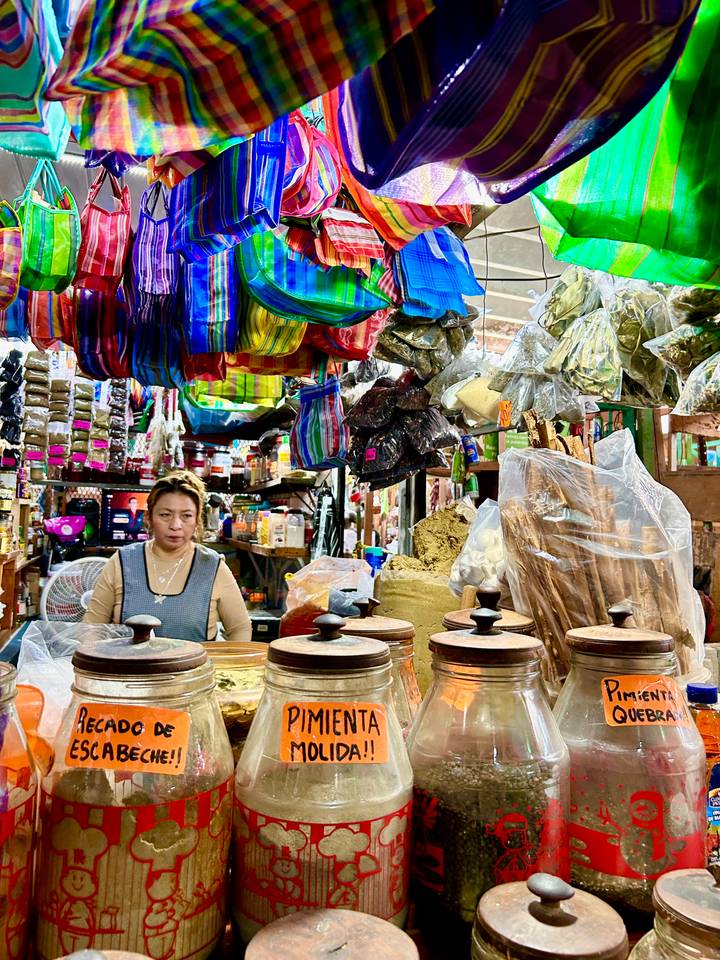 Market vendor surrounded by colorful hanging textiles and piles of spices inside a bustling bazaar.