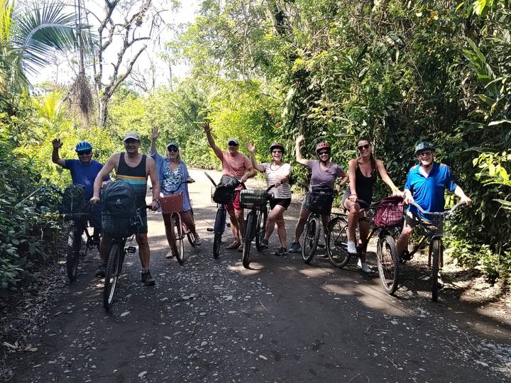 Cyclists pause for a photo on a shaded tropical dirt road surrounded by lush jungle vegetation.