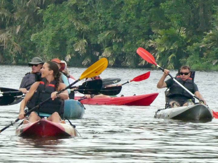 Several paddlers steer colorful kayaks together on a wide lagoon fringed by dense jungle.