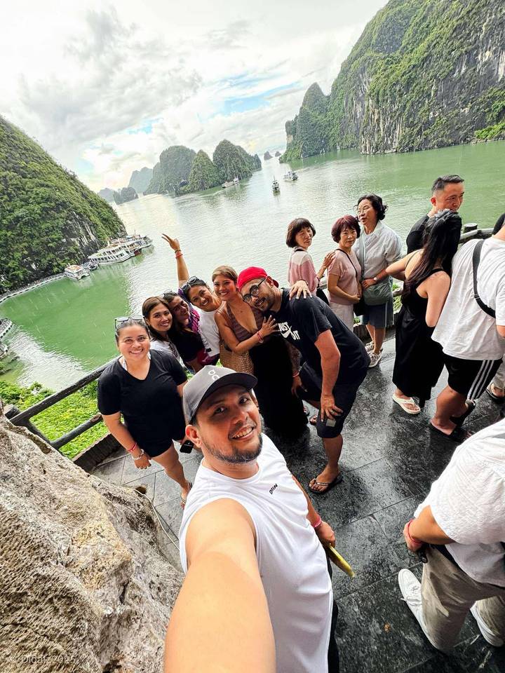 Group selfie on a cliffside lookout above emerald waters and cruise boats of Halong Bay.