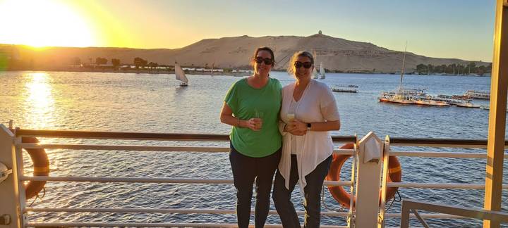 Two women enjoy drinks on a Nile cruise deck at golden sunset with feluccas sailing past sandy Aswan hills.