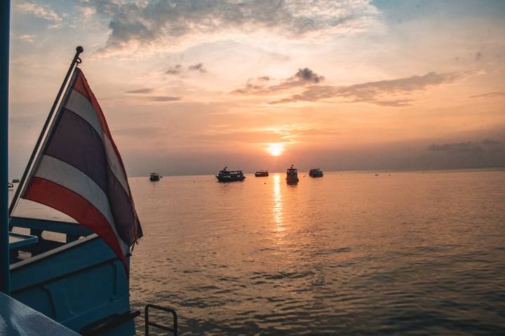 Thai flag flutters from a boat at sea as a warm sunset casts reflections over anchored vessels on calm waters.