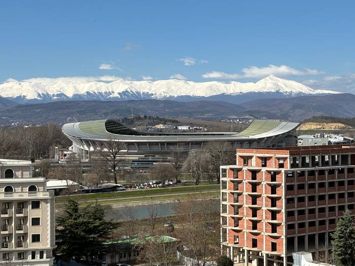 Modern oval stadium against river and snowy mountains on a clear day (duplicate view).