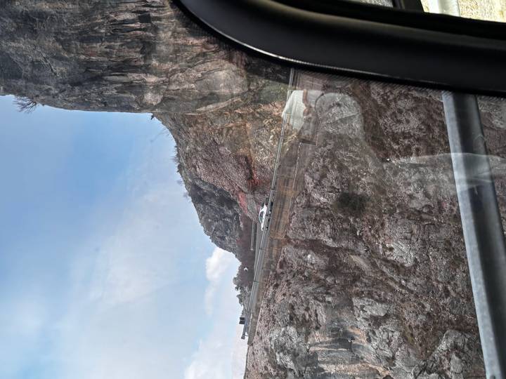 View through a vehicle window of a winding mountain road cut into steep rocky cliffs.