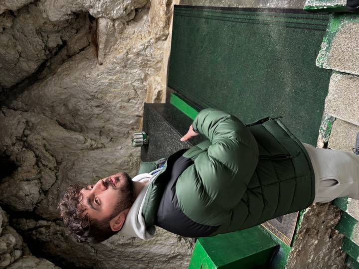 Tour guide gesturing toward a rock-walled shrine interior, wearing a green jacket and standing on a carpeted platform.