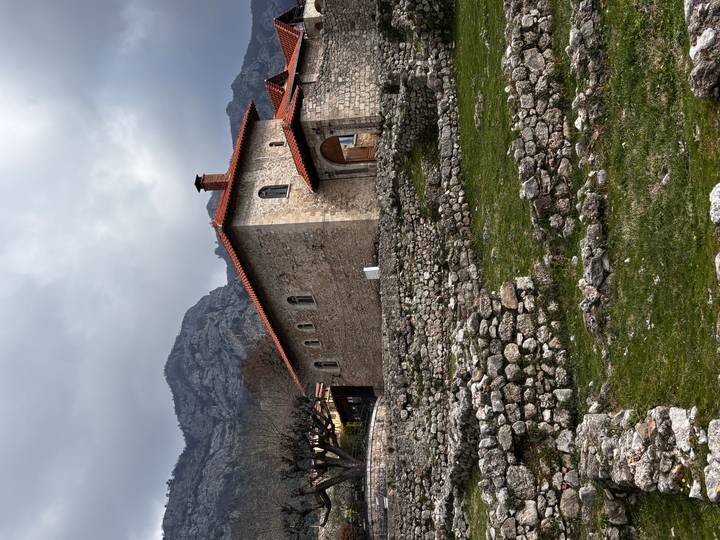 Stone church-like building with red-tiled roof perched on terraced ruins against a grey mountain backdrop.