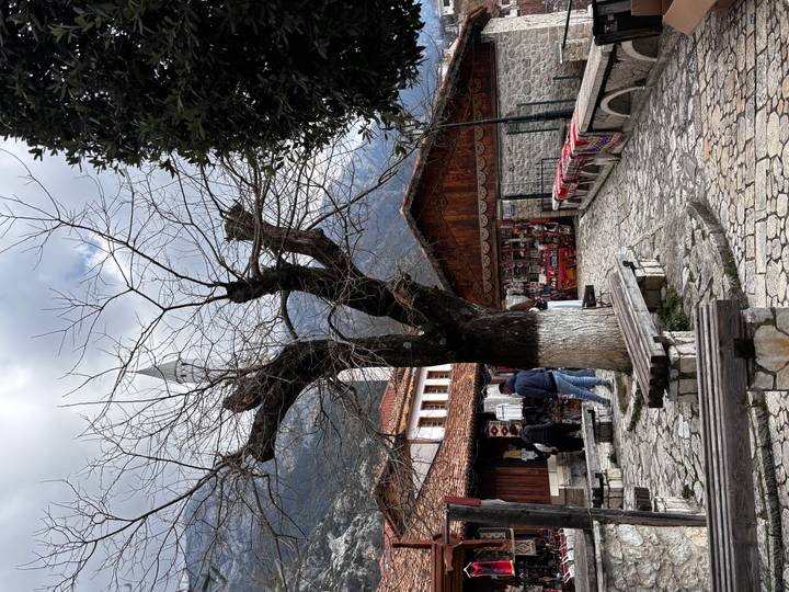 Cobblestoned village square with leafless tree, souvenir stalls and a mosque minaret rising behind.