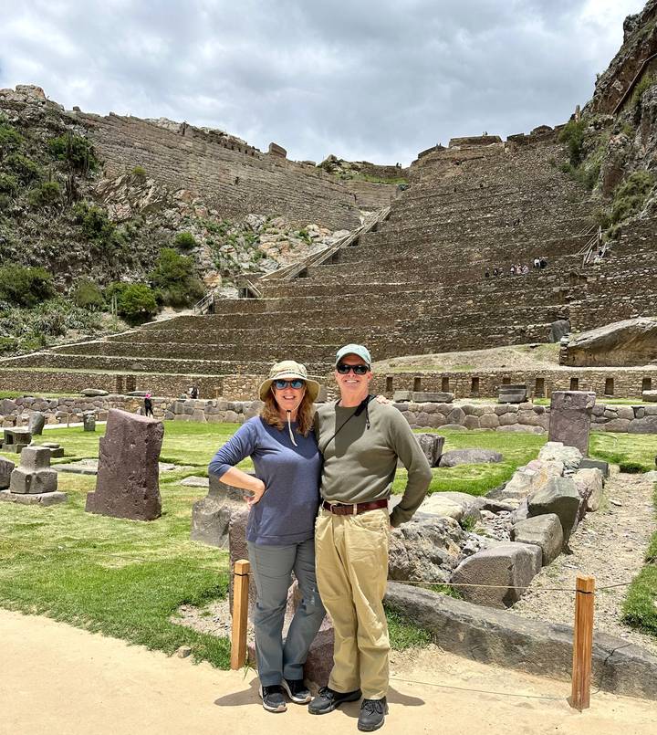A smiling couple poses before the terraced Inca ruins of Ollantaytambo under bright sun.