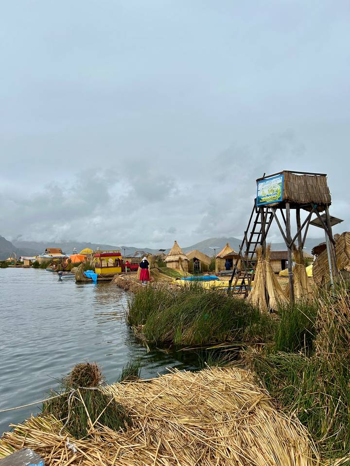 Reed huts and boats float on the Uros Islands while visitors explore under a grey sky.