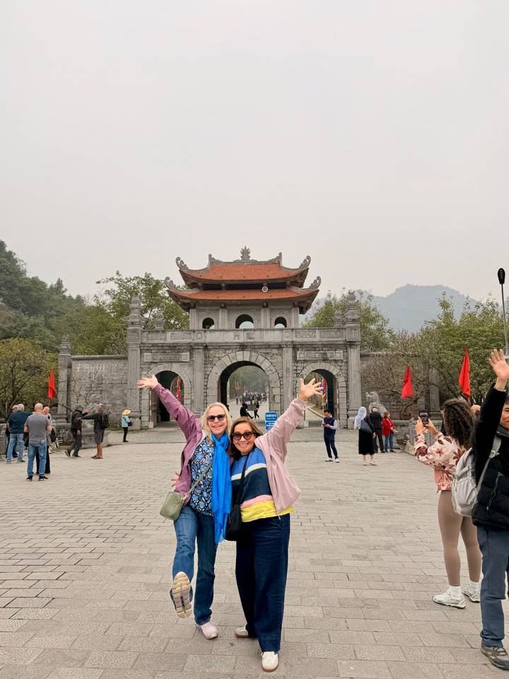 Tourists celebrate in front of a grand stone gate with three arches and pagoda roof in a misty valley.