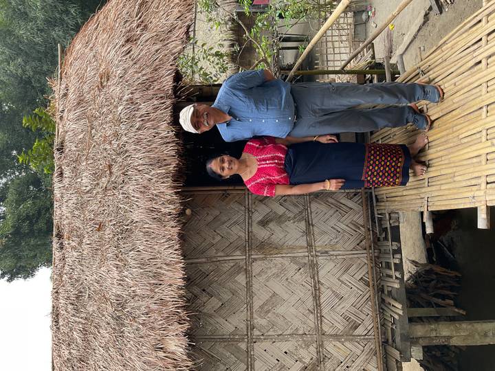 Smiling couple pose on a bamboo walkway beside a thatched hut in a rural village.