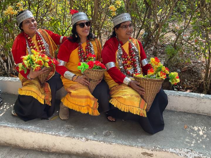 Three women in vibrant traditional Meghalayan attire sit holding baskets of colorful flowers.