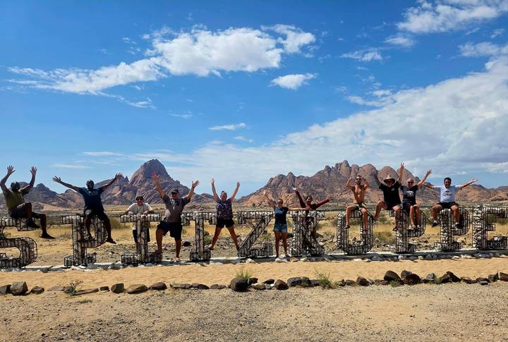 Excited tour group jumping in front of the metal Spitzkoppe sign with granite peaks behind.