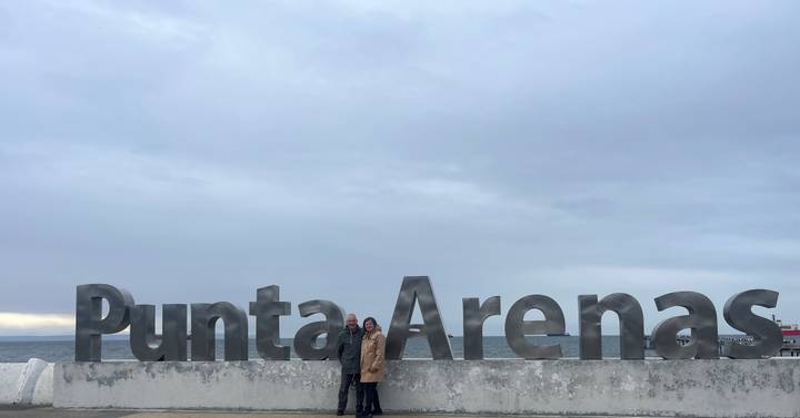 A couple poses in front of the large metallic Punta Arenas sign beside the sea under an overcast sky.