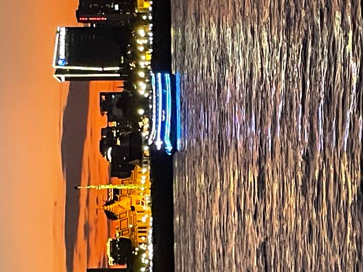 Blurry night shot of an illuminated boat on a river against an orange sunset sky and city lights.