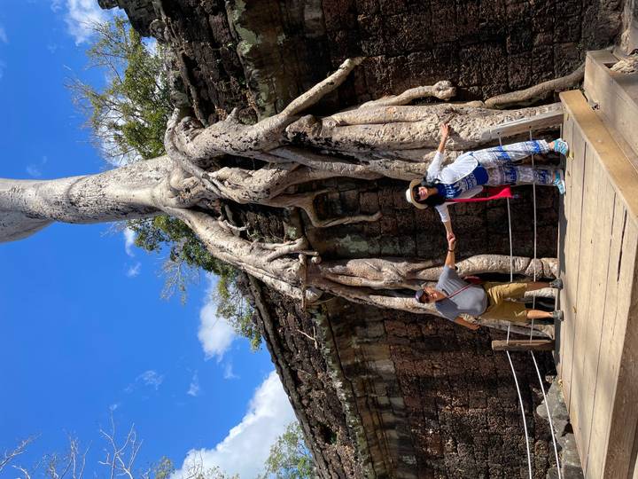 Couple playfully posing beneath enormous tree roots snaking over ancient stone temple walls under a sunny sky.