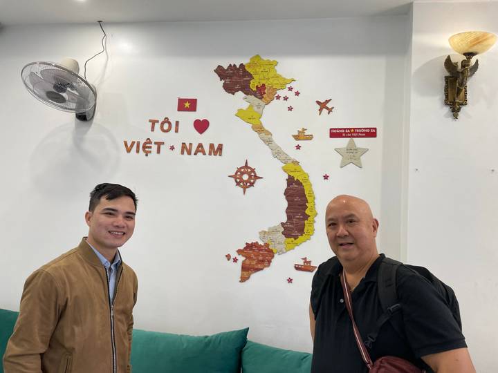 Indoor snapshot of two men standing beside a wall map of Vietnam decorated with icons and the national flag.