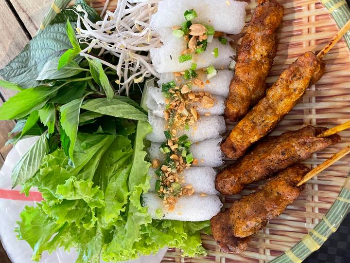 Vietnamese dish of grilled pork skewers with rice noodle rolls, crushed peanuts and fresh herbs on a bamboo tray.