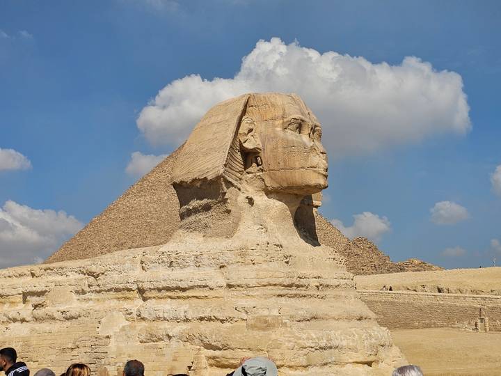 Close-up side view of the Great Sphinx with a pyramid rising behind it under a bright blue sky with scattered clouds.