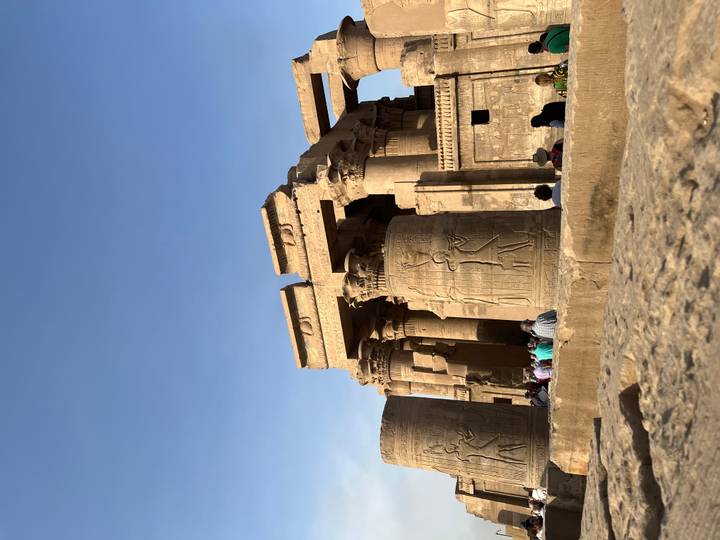 Massive sandstone columns and reliefs of Kom Ombo Temple with tourists gathered below.
