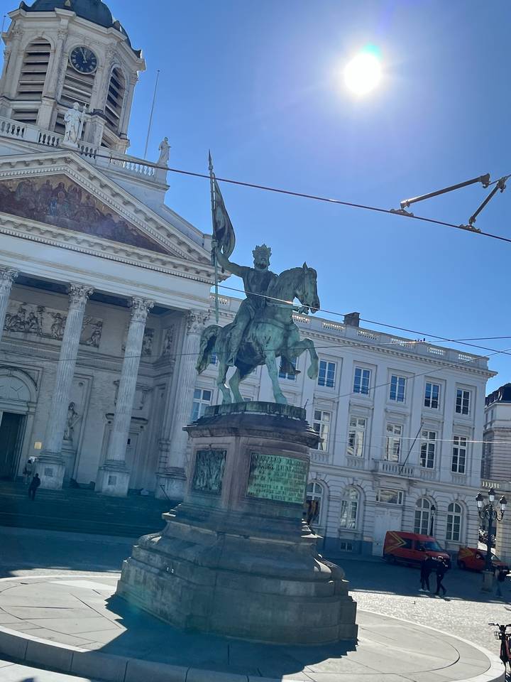 Bronze equestrian statue stands before a neoclassical building under a clear blue sky with tram wires overhead.