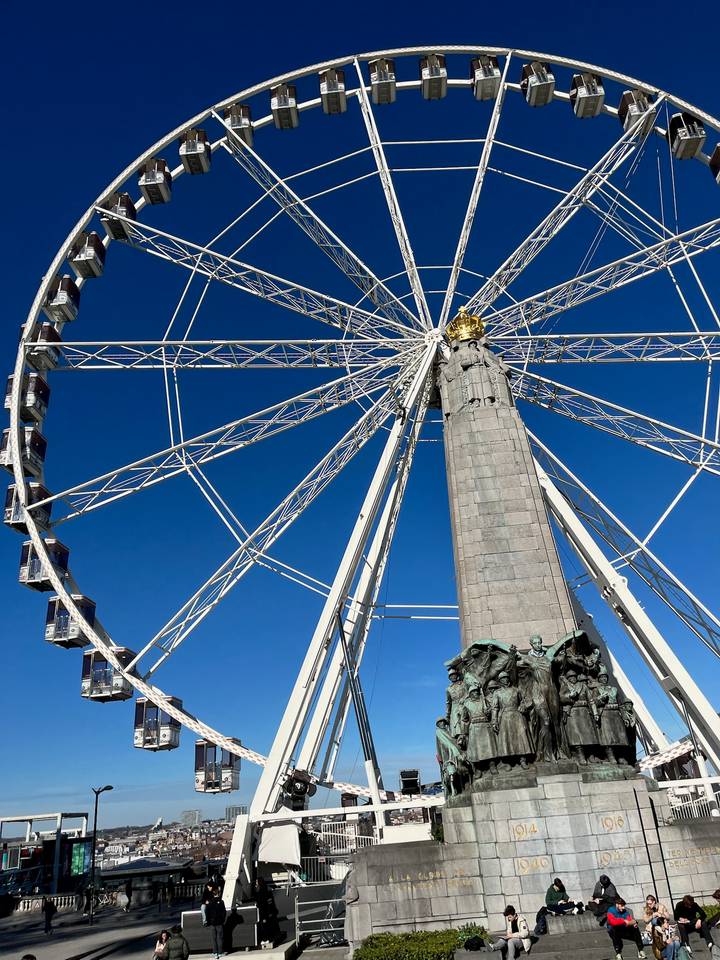 Upward view of a ferris wheel structure framing a tall column topped with a golden crown against deep blue sky.