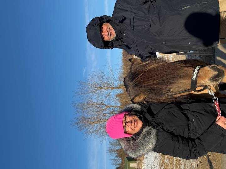 Smiling couple posing with an Icelandic horse on a sunny winter day with blue sky and bare trees.