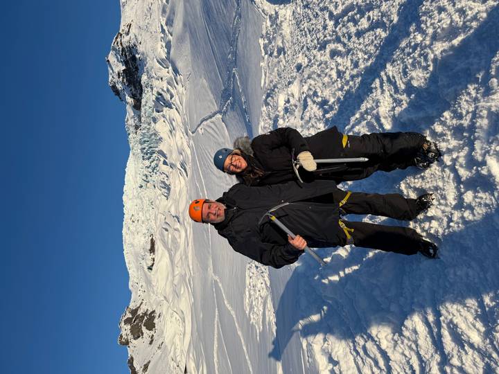 Two travellers in snow gear and helmets stand on a glacier with ice axes, bright sun illuminating the icy slope behind.