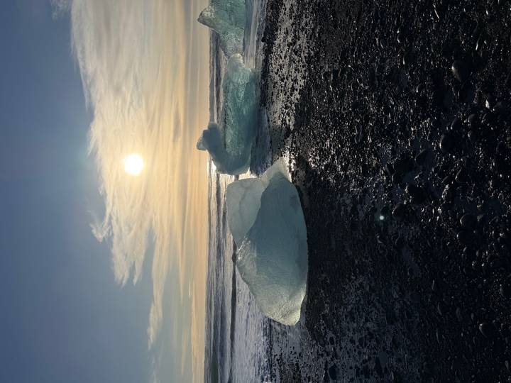 Translucent icebergs glitter on a black-pebble beach at sunrise with the sun low over the Atlantic waves.