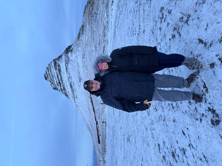 Couple in winter jackets standing on snowy ground with the conical Kirkjufell mountain rising behind them.