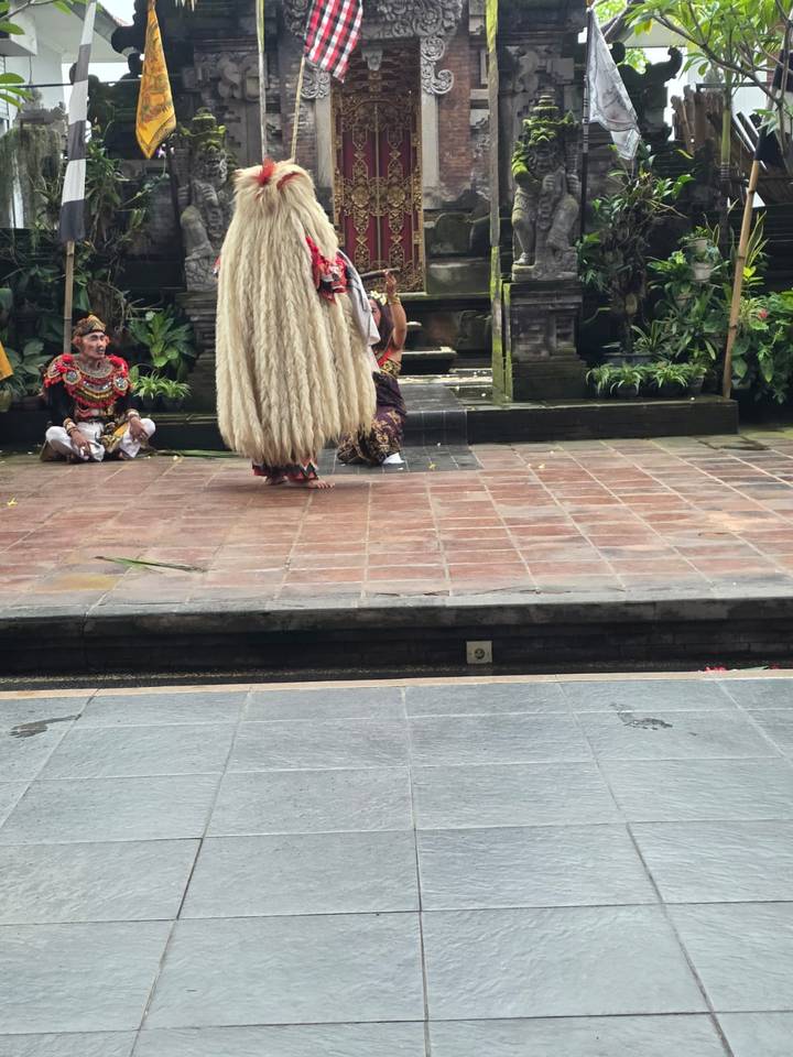 Barong performer with long white fur costume stands before seated actors on a tiled stage.
