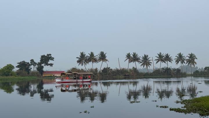 Traditional Kerala houseboat glides on calm backwaters lined with palm trees under a hazy sky.