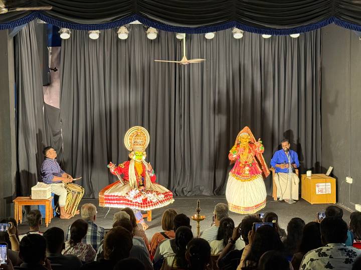 Kathakali dancers in elaborate costumes perform on a small theatre stage before an audience.