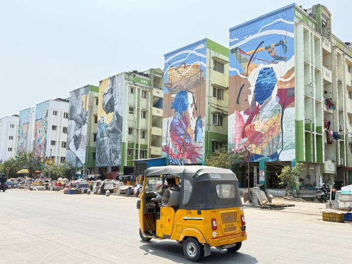 Street lined with high-rise buildings painted in bright murals as an auto-rickshaw drives by.