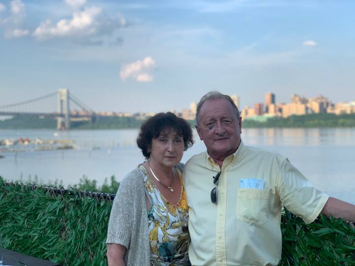 Older couple poses by a calm river with city skyline and suspension bridge behind.
