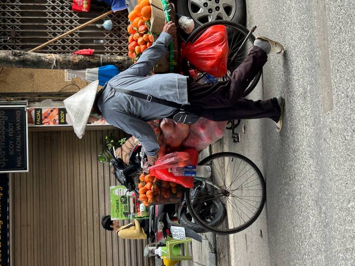 Street vendor pushing a bicycle loaded with bright oranges in mesh bags along a city road.