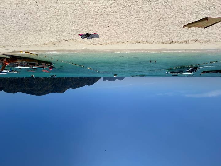 White-sand beach with long-tail boats and swimmers set before emerald sea and steep island cliffs.