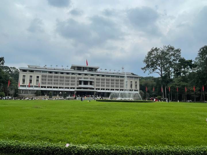 Wide lawn and fountain front the modernist Reunification Palace under partly cloudy skies.