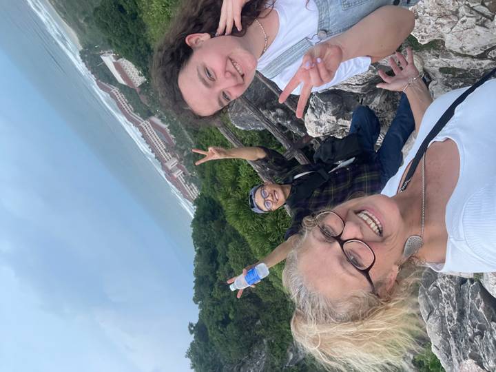 Three travellers flash peace signs atop a rocky lookout with ocean and resort buildings far below.