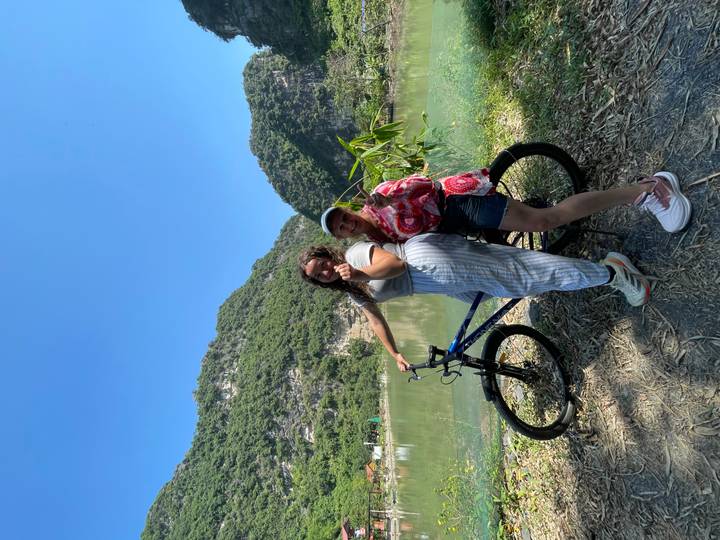 Two women with a bicycle pose beside a calm lake backed by dramatic limestone mountains under a clear blue sky.