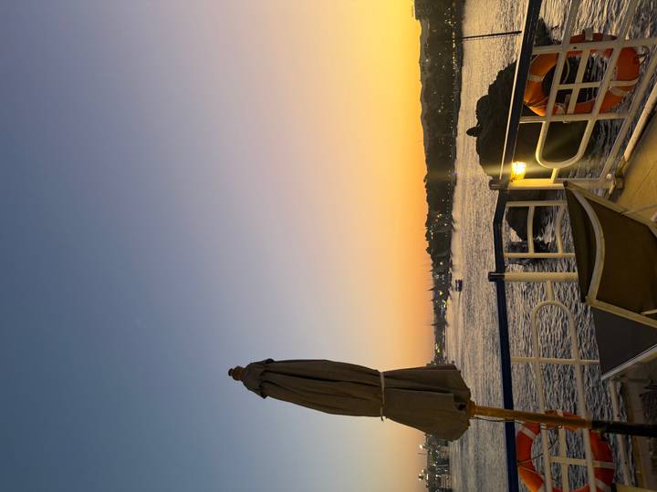 Deck chair and umbrella on a boat overlooking a river at colorful sunset with distant lights