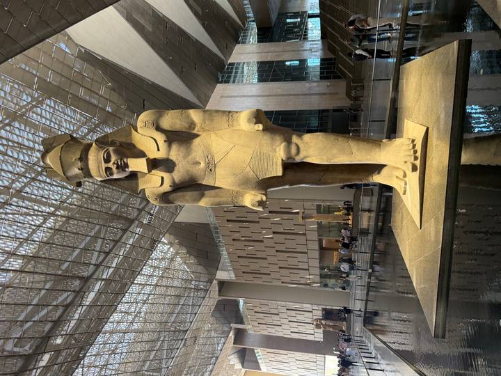 Colossal statue of a pharaoh standing in a grand modern museum atrium with visitors below.