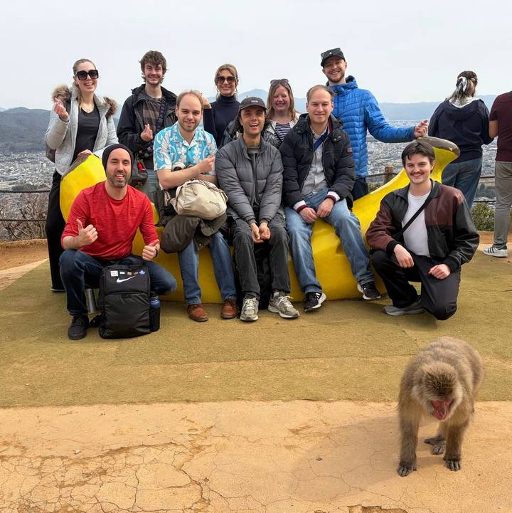Casual group photo around a large yellow banana sculpture at a hillside viewpoint with a curious monkey in the foreground.