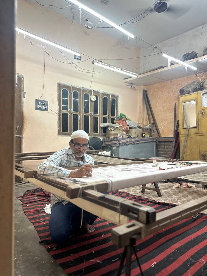 An artisan meticulously embroiders fabric on a wooden frame inside a cluttered workshop.