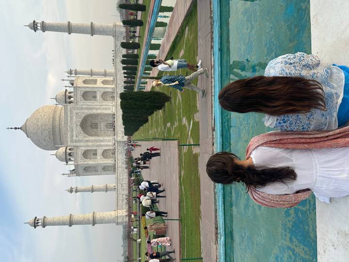 Visitors admire the Taj Mahal from its reflecting pool while two women sit foreground with backs turned.