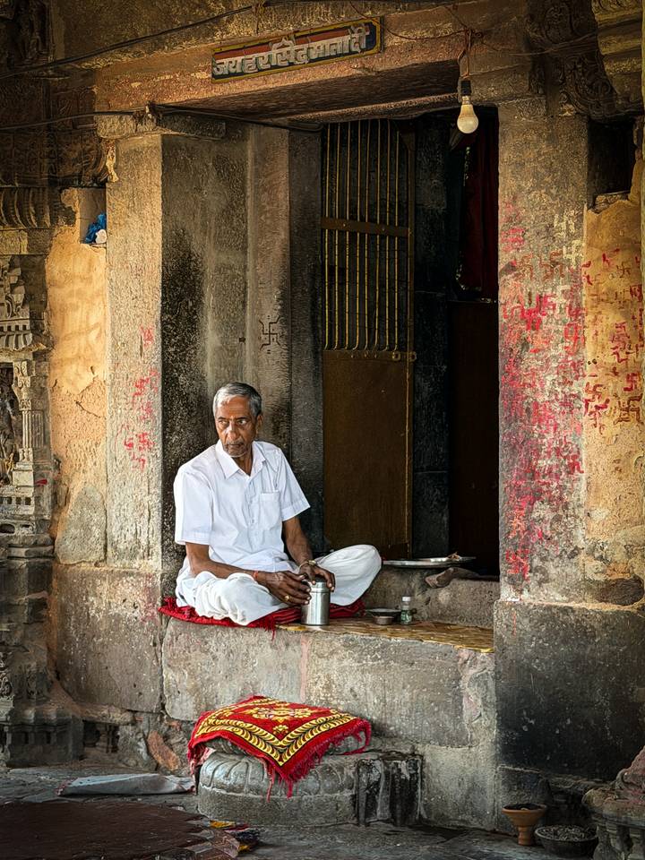 Elderly man in white clothing sits peacefully at the carved entrance of an Indian temple.